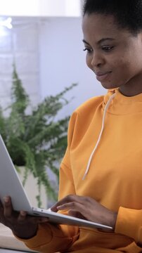 Young black woman in orange hoodie typing on laptop at home, online student lifestyle. remote work, freelance career, digital nomad