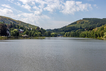 Fototapeta premium View of peaceful lake surrounded by lush green hills under a bright cloudy sky at Lac des Doyards - Belgium