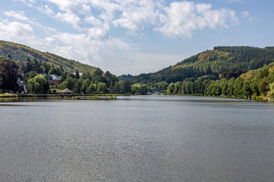 View of peaceful lake surrounded by lush green hills under a bright cloudy sky at Lac des Doyards - Belgium