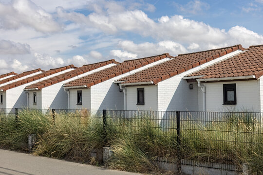 Row of identical white bungalows with red tiled roofs behind a metal fence and tall dune grass in Westende - Belgium