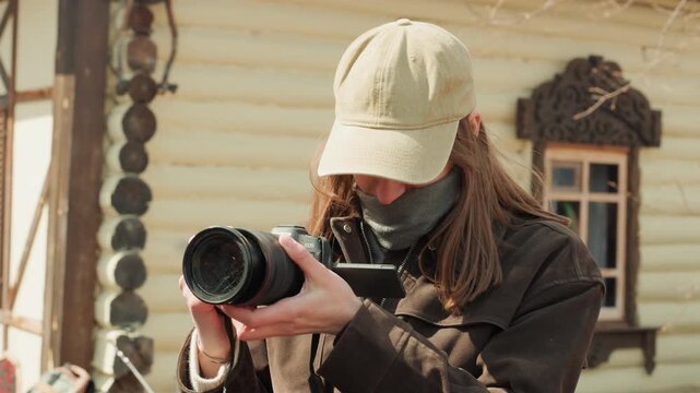 Young woman in cap and face mask carefully adjusts camera lens beside rustic wooden building, leaning forward with both hands on equipment and calm focused posture in warm daylight