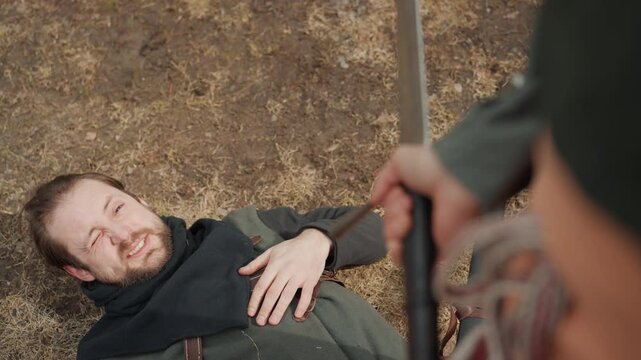 Overhead scene shows standing figure holding polearm upright beside fallen person on brown soil, with hand on chest and muted natural light creating tense aftermath and dramatic uncertainty