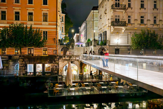 The Ribja brv Fish Footbridge crossing the Ljubljanica River in the old town of Ljubljana, Slovenia, looking toward the floating Makalonca terrace bar and cafe area along the embankment at night.