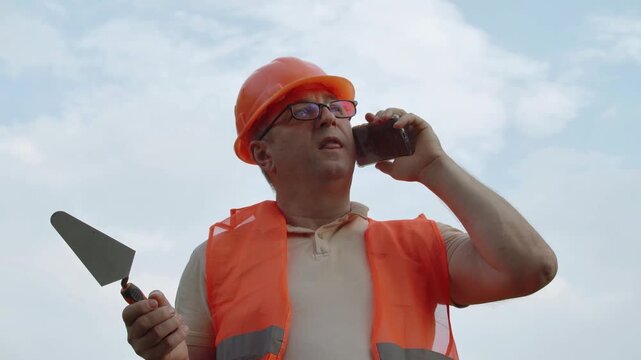 Construction worker in orange safety vest and hard hat standing outside, holding smartphone to ear and trowel in other hand, against cloudy sky. Foreman giving instructions.