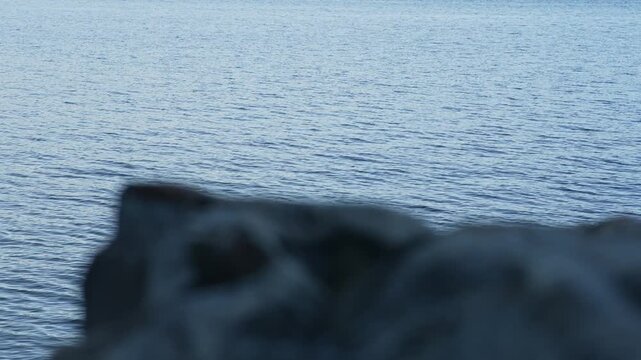 Rippling lake water appears beyond blurred rocks in foreground, creating a shallow depth of field composition with soft blue water texture and dark out of focus stones along the shoreline