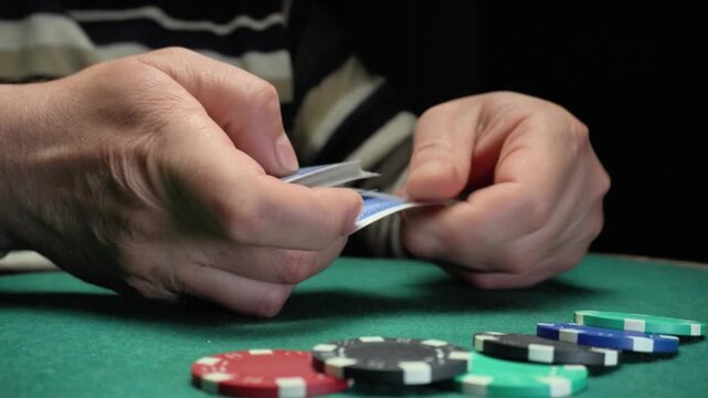 Hands of a person with light skin shuffle a deck of playing cards while seated at a green felt poker table with colorful poker chips arranged in a row in the foreground