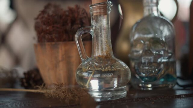 Clear glass decanter with handle and short spout rests on weathered wooden tabletop beside decorative glass skull bottle and potted dried plant; scene shows no visible motion and emphasizes still