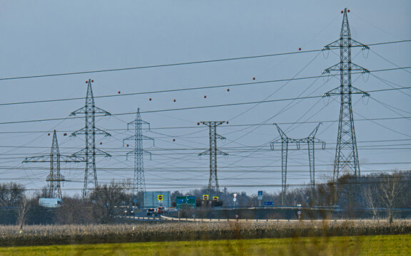 Dense network of high-voltage transmission lines and electricity pylons beside a road junction and agricultural landscape, illustrating modern power and transport infrastructure.