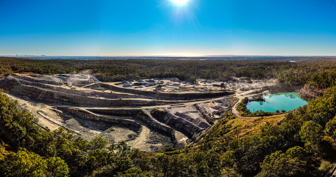 Aerial view of Holcim Australia - Petrie Quarry featuring an open-pit mine with a turquoise reservoir surrounded by dense forest under a clear blue sky in Kurwongbah, Queensland, Australia.