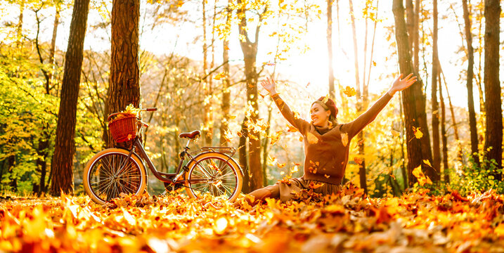Woman is having fun in an autumn park, overlooking bicycle at sunset. Young woman throws bright yellow leaves while walking through sunny park. Concepts of relaxation, walking, and seasonality.