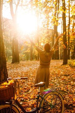 Woman is having fun in an autumn park, overlooking bicycle at sunset. Young woman throws bright yellow leaves while walking through sunny park. Concepts of relaxation, walking, and seasonality.