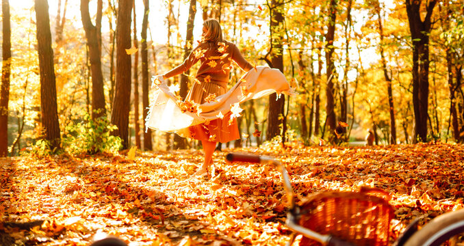 Woman is having fun in an autumn park, overlooking bicycle at sunset. Young woman throws bright yellow leaves while walking through sunny park. Concepts of relaxation, walking, and seasonality.