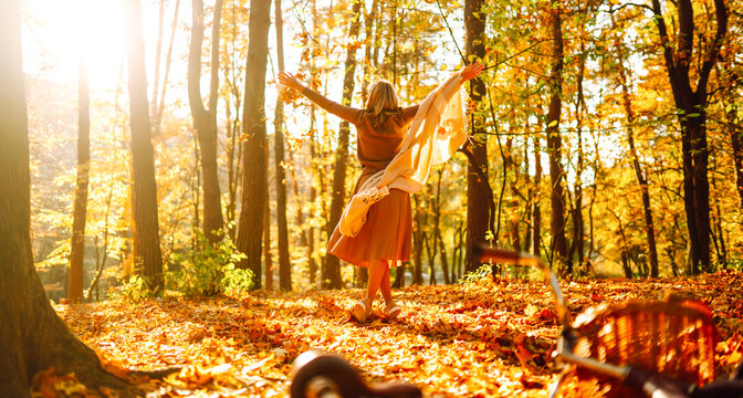 Woman is having fun in an autumn park, overlooking bicycle at sunset. Young woman throws bright yellow leaves while walking through sunny park. Concepts of relaxation, walking, and seasonality.