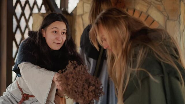 Woman offers dried flower bundle while smiling and reaching toward companion near warm wood-fired oven; visible actions include handing, receiving, turning, and open interaction inside cozy rustic