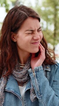 Young woman having toothache walking in city street touches cheek with dental discomfort. Lady tourist feeling sick outdoors suffers sudden tooth pain during urban trip near park needing dentist care.