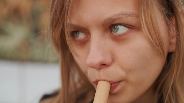 Young woman with freckled skin gently holding biscuit between lips, chewing softly while looking aside with thoughtful calm, warm diffused daylight and soft blurred background, with woman, young