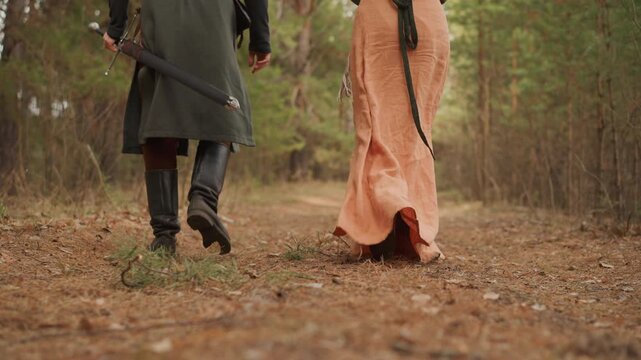 Low rear viewpoint shows two people walking forward along leaf-covered woodland trail, with lifted heels, flowing hems, soft dappled light, and quiet natural surroundings suggesting calm