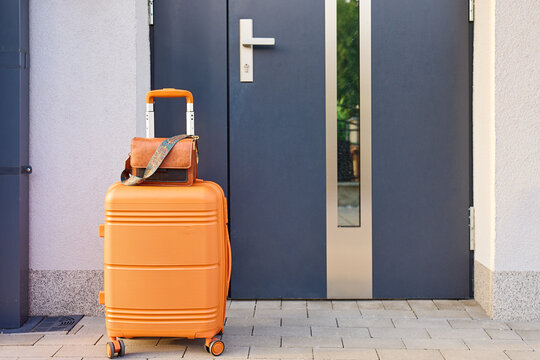 Orange rolling suitcase with handbag stands outside a front door