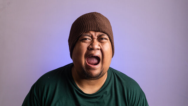 A young Southeast Asian male shouts loudly with a wide open mouth and a deeply expressive face against a vibrant purple background. The adult individual wears a brown knitted beanie