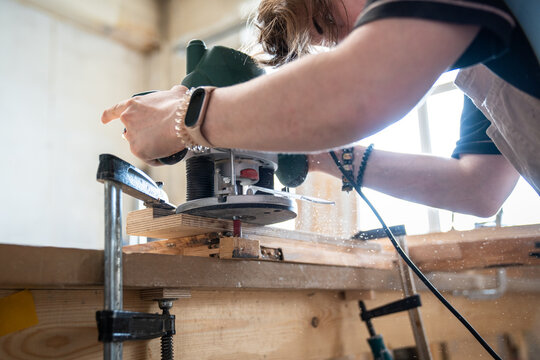 Skilled woman carpenter working with electric wood router machine at workplace in woodworking studio, craftswoman processing wood with handheld plunge router, milling machine trims and cuts surface.