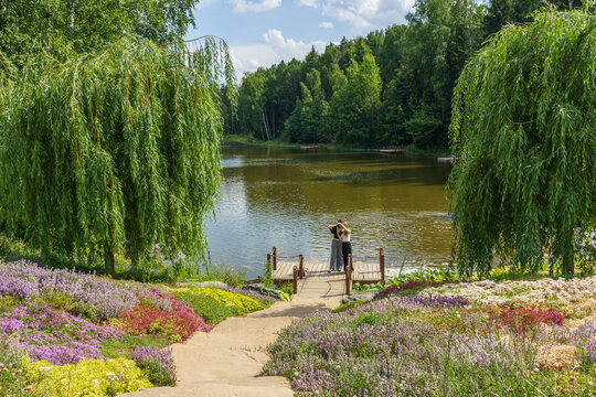 Woman in black dress make selfy on wooden dock by river surrounded by lush greenery and colorful flowers under a bright blue sky with fluffy clouds