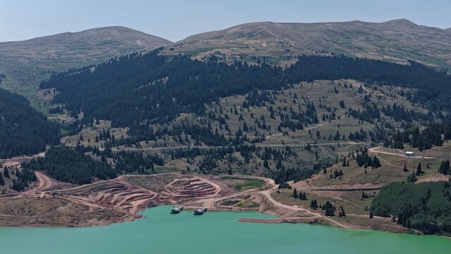 Aerial view of Mayflower Tailings Pond with its turquoise water surrounded by coniferous forests, rugged mountain slopes, and dirt access roads in Summit County, Colorado, United States.