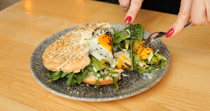 Diner presses fork into greens cutting sunny egg near sesame bun. Yolk flows over arugula and person gathers tasty bite while keeping plate steady in cafe
