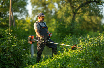 A gardener stands on a slope, operating a string trimmer to cut grass and weeds. The sun shines through trees, making the scene bright and active in the afternoon © Віталій Б.