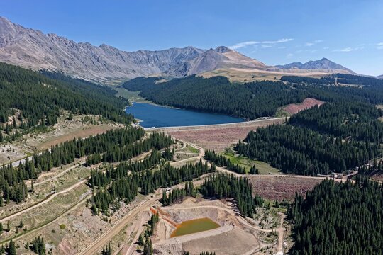 Aerial view of Clinton Gulch Dam Reservoir surrounded by dense pine forests and rugged mountains under a clear blue sky in Summit County, Colorado, United States.