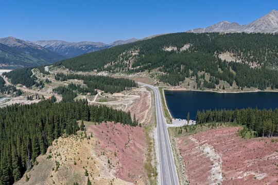 Aerial view of Clinton Gulch Dam Reservoir surrounded by dense pine forests, rugged mountain terrain, and a winding highway in Summit County, Colorado, United States.