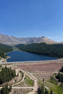 Aerial view of Clinton Gulch Dam Reservoir surrounded by dense pine forests and rugged mountains under a clear blue sky in Summit County, Colorado, United States.