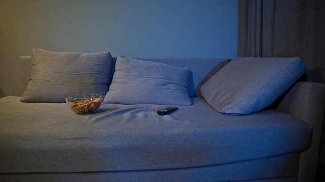 Empty sofa glows beneath flickering television light in a dark living room, with a bowl of snacks and TV console nearby. A quiet domestic scene suggesting solitude, absence, passive entertainment.