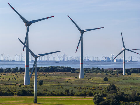 Aerial view of wind turbines standing in green fields near the coast with a distant industrial skyline in Emden, Niedersachsen, Germany.