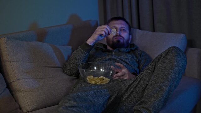 Exhausted man lounges on a couch under the glow of a television, snacking and drifting toward sleep while holding a remote, portraying sedentary habits, passive media consumption, boredom, isolation.