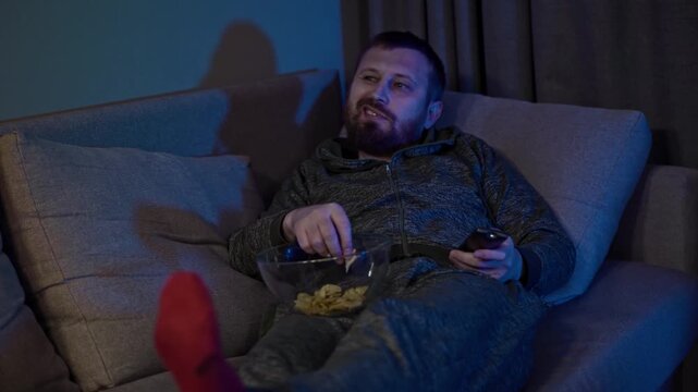 Tired man relaxes alone on a sofa as changing television light illuminates his face, gradually falling asleep with a bowl of chips nearby, portraying passive entertainment, screen addiction.