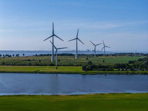 Aerial view of wind turbines standing in a green field next to a river with a distant industrial skyline in Emden, Niedersachsen, Germany.