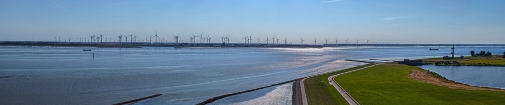 Aerial view of the Volkswagenwerk Emden factory and wind turbines along the Ems river estuary, with green dikes and calm water under a clear blue sky in Emden, Niedersachsen, Germany.