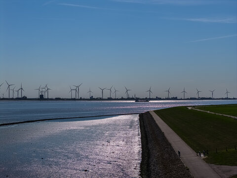 Aerial view of a wind farm with numerous turbines standing along the coastline, featuring a paved dike path in the foreground and calm water reflecting the sky in Emden, Niedersachsen, Germany.