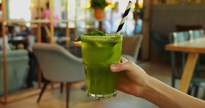 Female hand holds tall green smoothie with mint leaves and striped straw against cafe furniture. Drink appears near chairs waiting for refreshing sip at break
