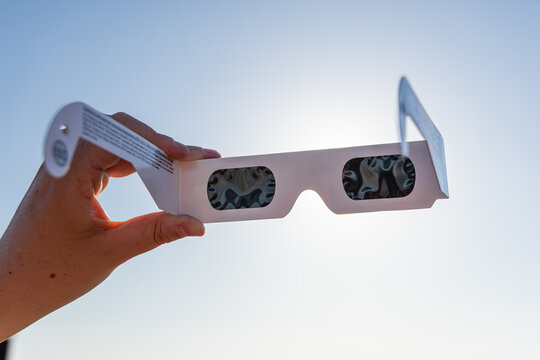 Gros plan sur une paire de lunettes d&rsquo;&eacute;clipse solaire, avec grands verres filtrants noirs pour observer le soleil. En arri&egrave;re plan, le soleil avec un ciel bleu.