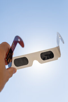 Gros plan sur une paire de lunettes d&rsquo;&eacute;clipse solaire, avec grands verres filtrants noirs pour observer le soleil. En arri&egrave;re plan, le soleil avec un ciel bleu. Le format est vertical.