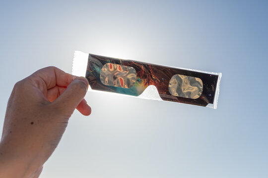 Une main tenant une paire de lunettes d&rsquo;&eacute;clipse solaire encore sous sachet plastique, neuve. En arri&egrave;re plan, le soleil avec un ciel bleu.
