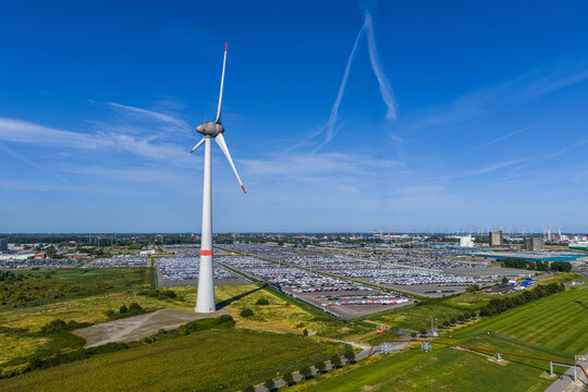 Aerial view of the Volkswagenwerk Emden factory with a large wind turbine in the foreground and rows of parked cars under a clear blue sky in Emden, Niedersachsen, Germany.