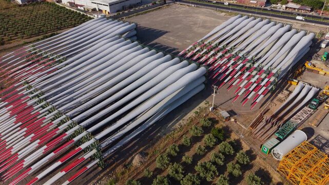 Aerial view of a wind turbine blade storage and manufacturing facility with dozens of enormous composite rotor blades arranged in rows, ready for transport and wind farm installation.