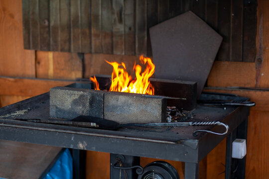 Fire blazes in a metal forge set on a workbench, surrounded by bricks and tools against a wooden wall backdrop