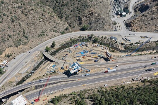 Aerial view of the highway construction site with cranes, heavy machinery, and bridge work in progress near the river in the mountainous terrain of Floyd Hill, Colorado, United States.