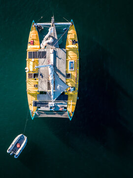 Aerial view of a yellow catamaran sailing boat with a small dinghy attached, floating on calm dark blue water in Cascais, Lisbon, Portugal.