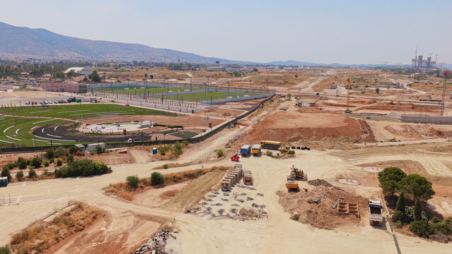 Aerial view of a large construction site featuring heavy machinery, earthworks, and green sports fields in the background in Athens, Greece.
