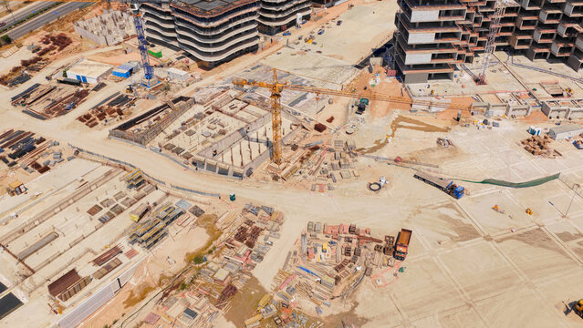 Aerial view of a large construction site featuring multiple cranes, building foundations, stacked construction materials, and partially completed modern apartment buildings in Athens, Greece.