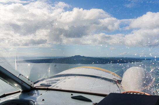 A cockpit‑view photograph taken from a small seaplane in Auckland, New Zealand, looking out across calm harbour water toward Rangitoto Island&rsquo;s volcanic peak under a partly cloudy sky